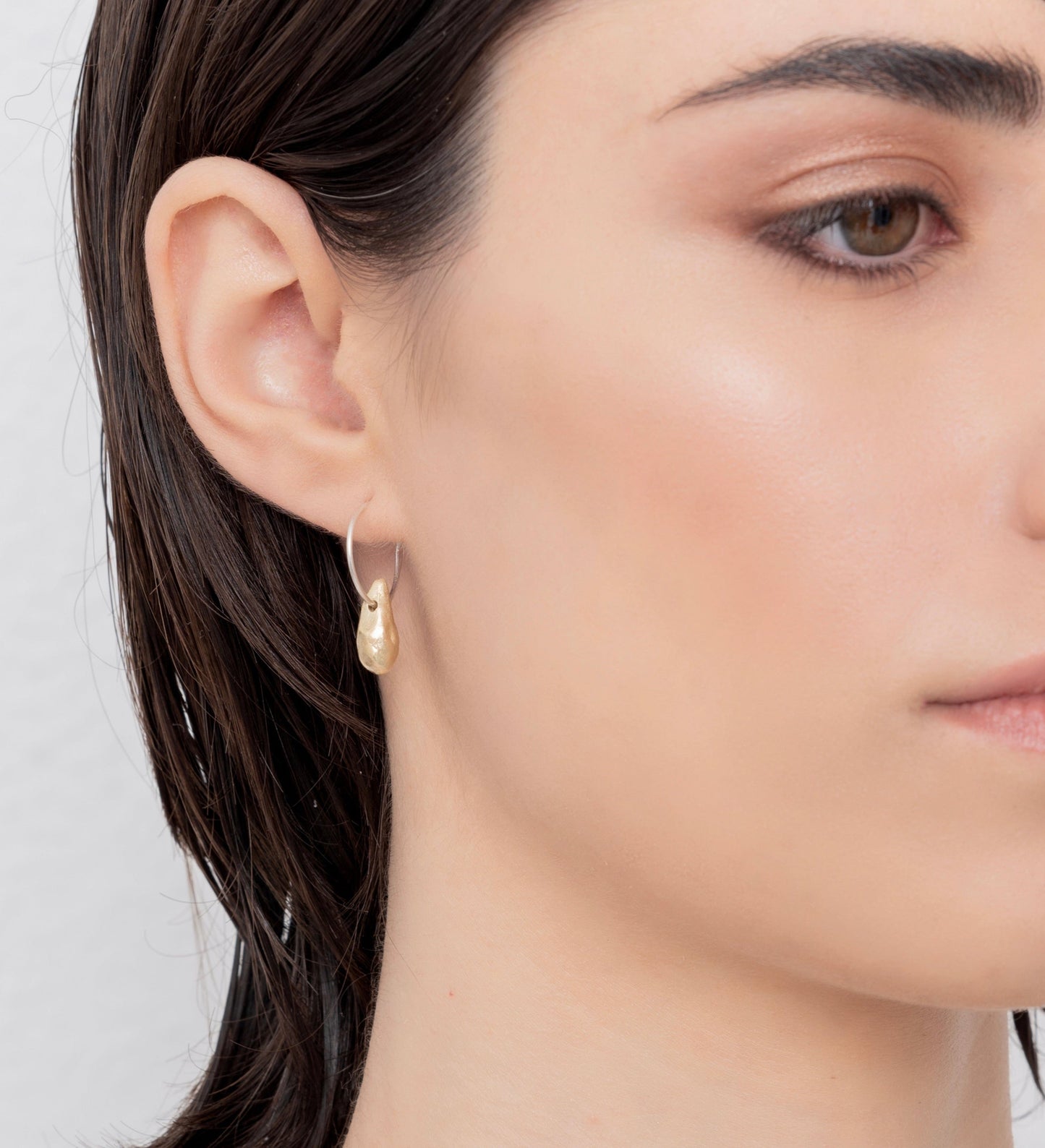 Close-up of a person with short, dark hair wearing Golden Drop Hoop Earrings Nautical Inspired, handmade by Angela Lago, looking slightly to the side with a neutral expression.