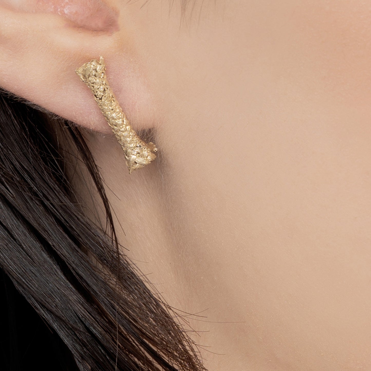 Close-up of a person's ear wearing the Sterling Silver Rope S Earring: Handmade Nautical Jewel, featuring a textured, short, irregular bar design with a gold-plated finish. The person's dark hair is partially visible.