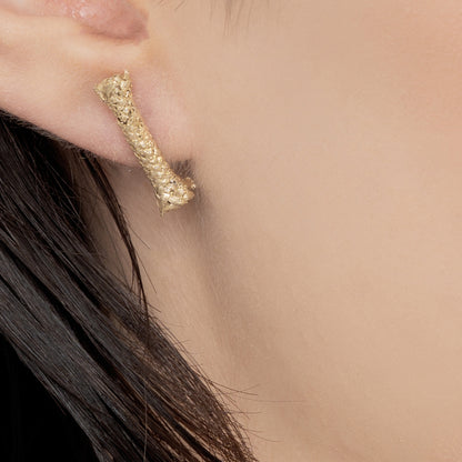 Close-up of a person's ear wearing the Sterling Silver Rope S Earring: Handmade Nautical Jewel, featuring a textured, short, irregular bar design with a gold-plated finish. The person's dark hair is partially visible.