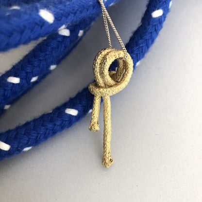 A Nautical golden knot pendant - Ballestrinque S, in the shape of a Clove Hitch knot, hangs in front of a blue and white polka-dotted braided rope.