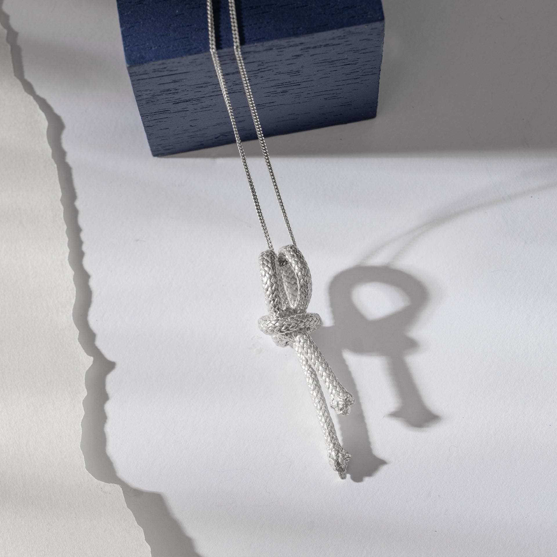 A Ballestrinque Small Nautical Knot Pendant in Sterling Silver, hanging over a blue wooden block, casts a heart-shaped shadow on a white surface.