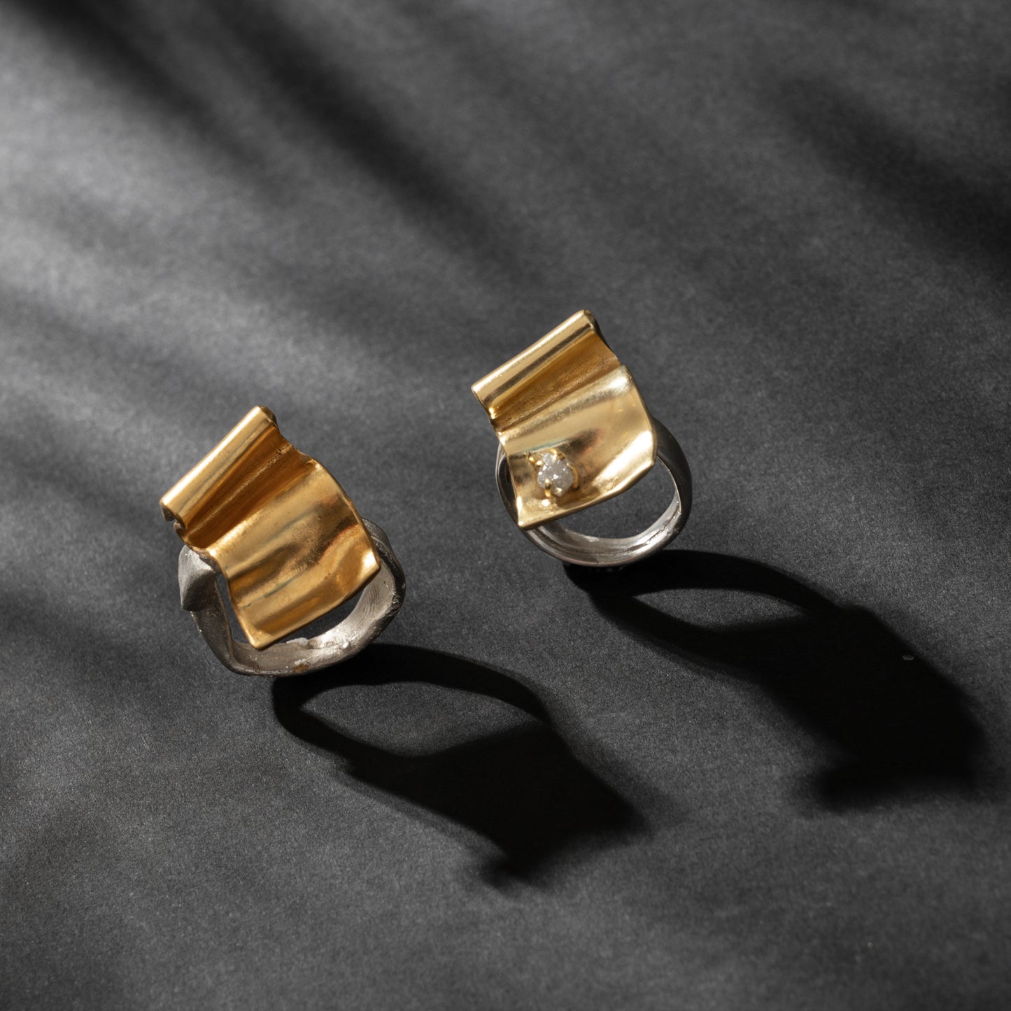 Two modern rings, one a Handcrafted Ring + Wave Gold 24k Plated Brass and the other made from sterling silver with abstract designs, featuring a small gemstone, displayed on a dark, shadowed background.