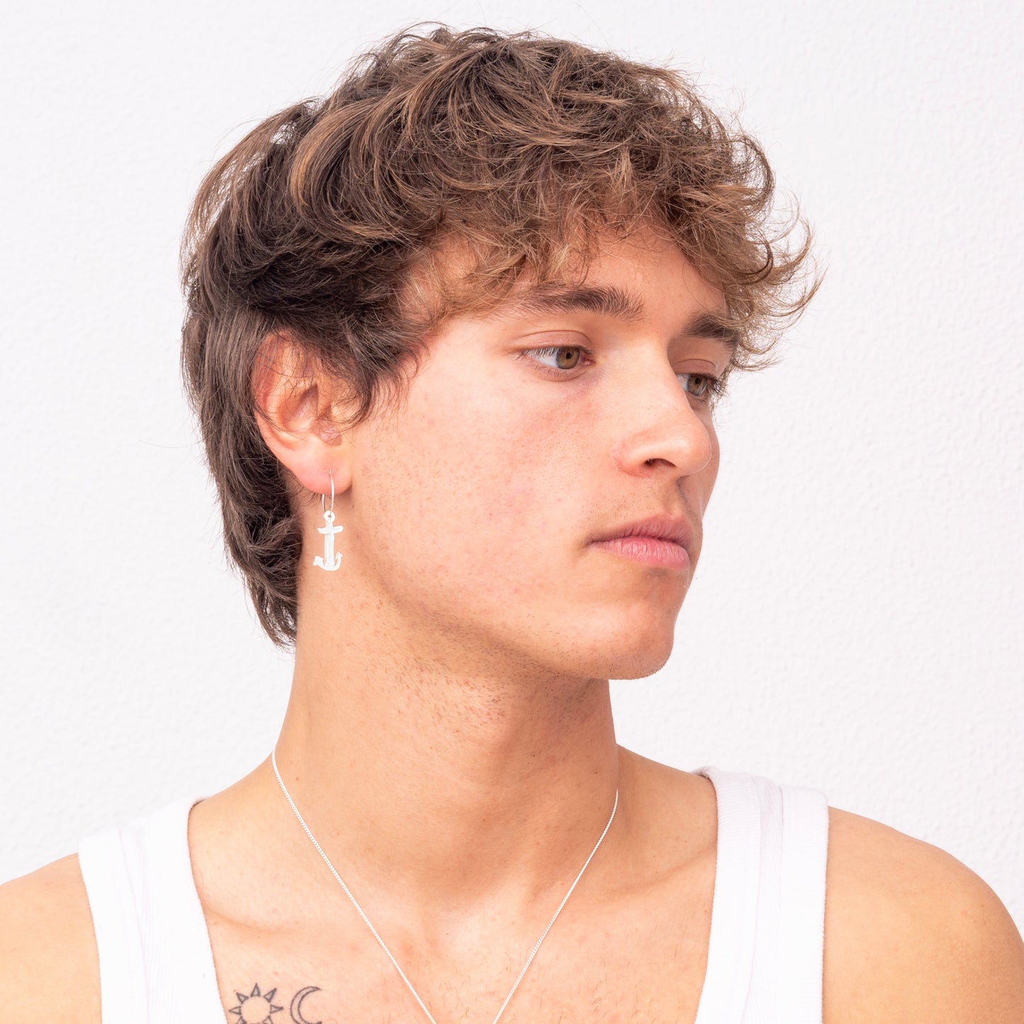 A person with wavy brown hair wearing a white tank top, an Underwater Anchor Handmade Nautical Hoop Earring by Angela Lago, and a necklace.