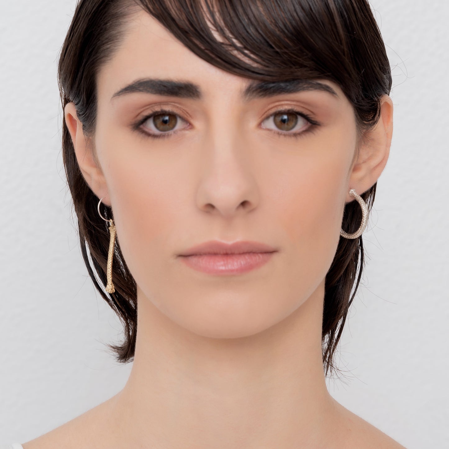 A person with short, dark hair is facing forward. They are wearing Nautical Hoop Corto Maltese Inspired Creole Earrings, made of sterling silver and featuring a rope design in size M, and they have a neutral expression. The background is plain and light-colored.