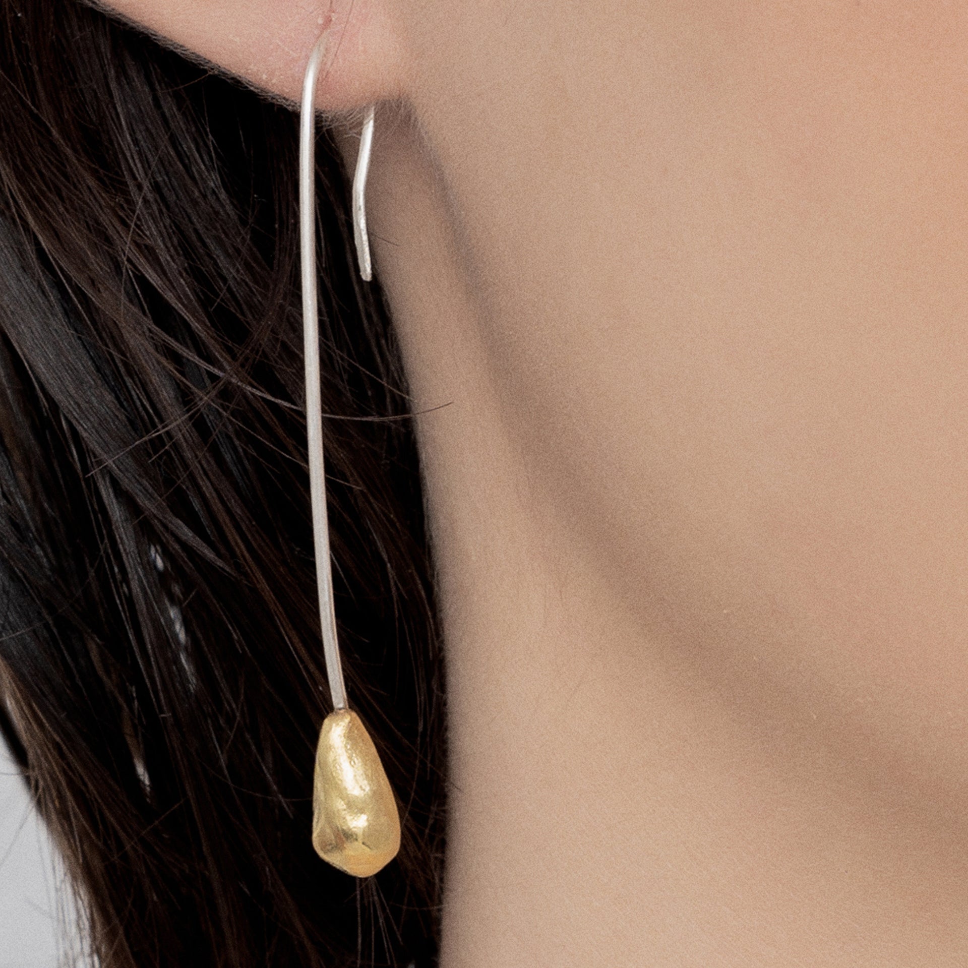 Close-up of a person wearing the Golden Drop Long Earring Handmade, featuring a sterling silver base with a dangling 24k gold plated teardrop-shaped charm meticulously crafted by Angela Lago.