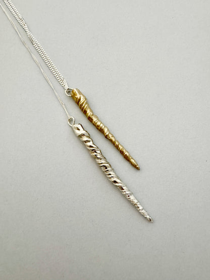 Two twisted metal pendants, one silver and one gold, dangle from delicate chains against a plain gray background. This exquisite Narwhal Tusk Handmade Tooth Pendant is crafted by Angela Lago, highlighting her intricate craftsmanship.