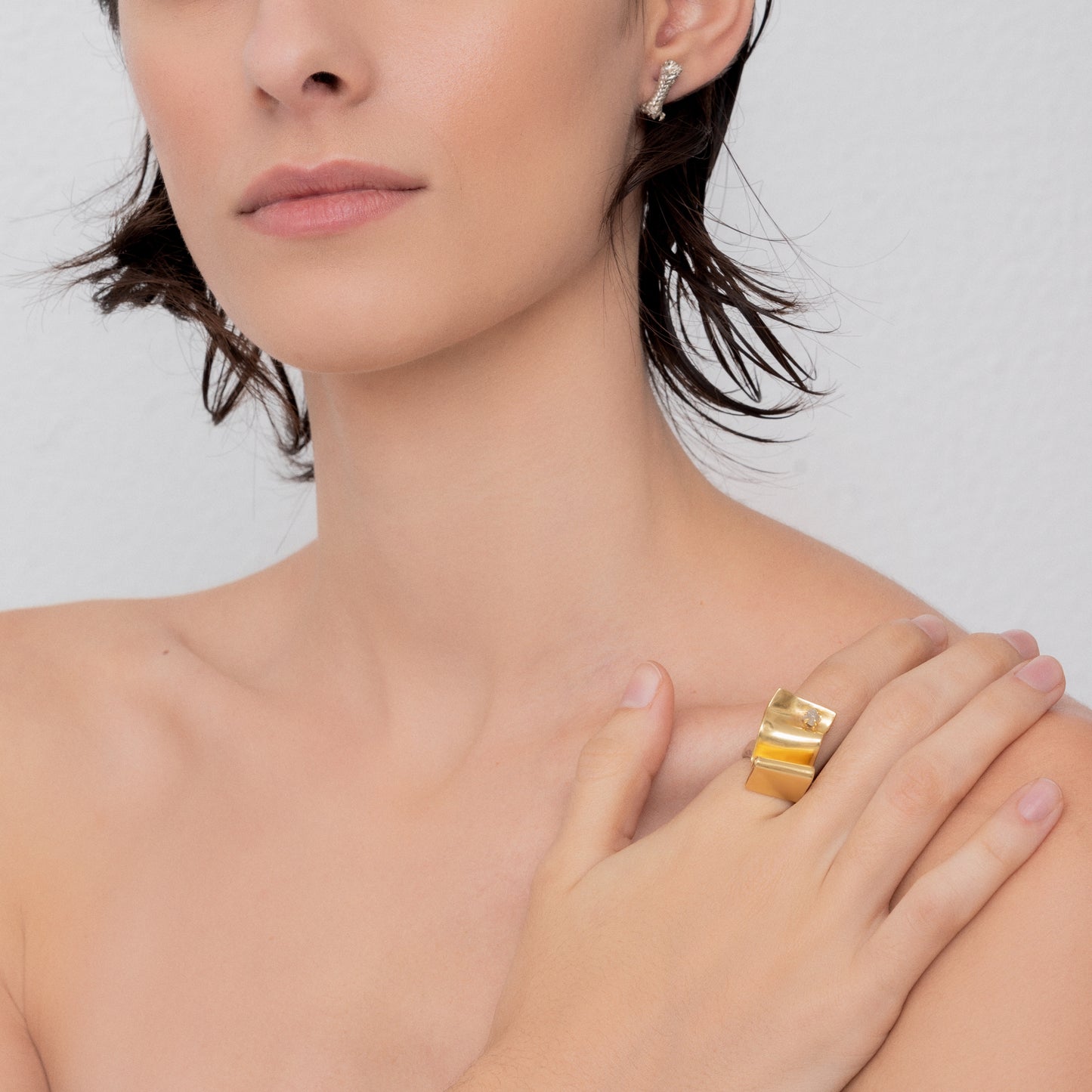 A woman with short brown hair and light skin, wearing the Golden Handcraft Ring Wave with Raw Diamond and a silver ear cuff, showcases her left hand adorned with a sterling silver ring on her shoulder against a plain background.