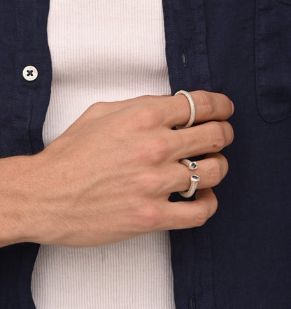 Close-up of a hand wearing a wedding ring and the Just a Nautical Handcrafted Rope Ring Inspired by Sailors, featuring a dark blue stone, against a white shirt and navy blue button-up shirt.