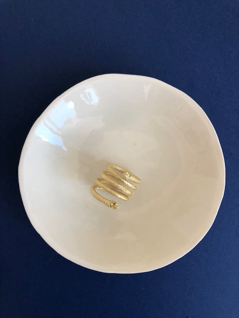 A Curly Handcrafted Rope Ring III, meticulously gold-plated brass (5 microns gold), is elegantly placed in the center of a white ceramic bowl against a deep blue background, showcasing Angela Lago's exquisite handmade craftsmanship.