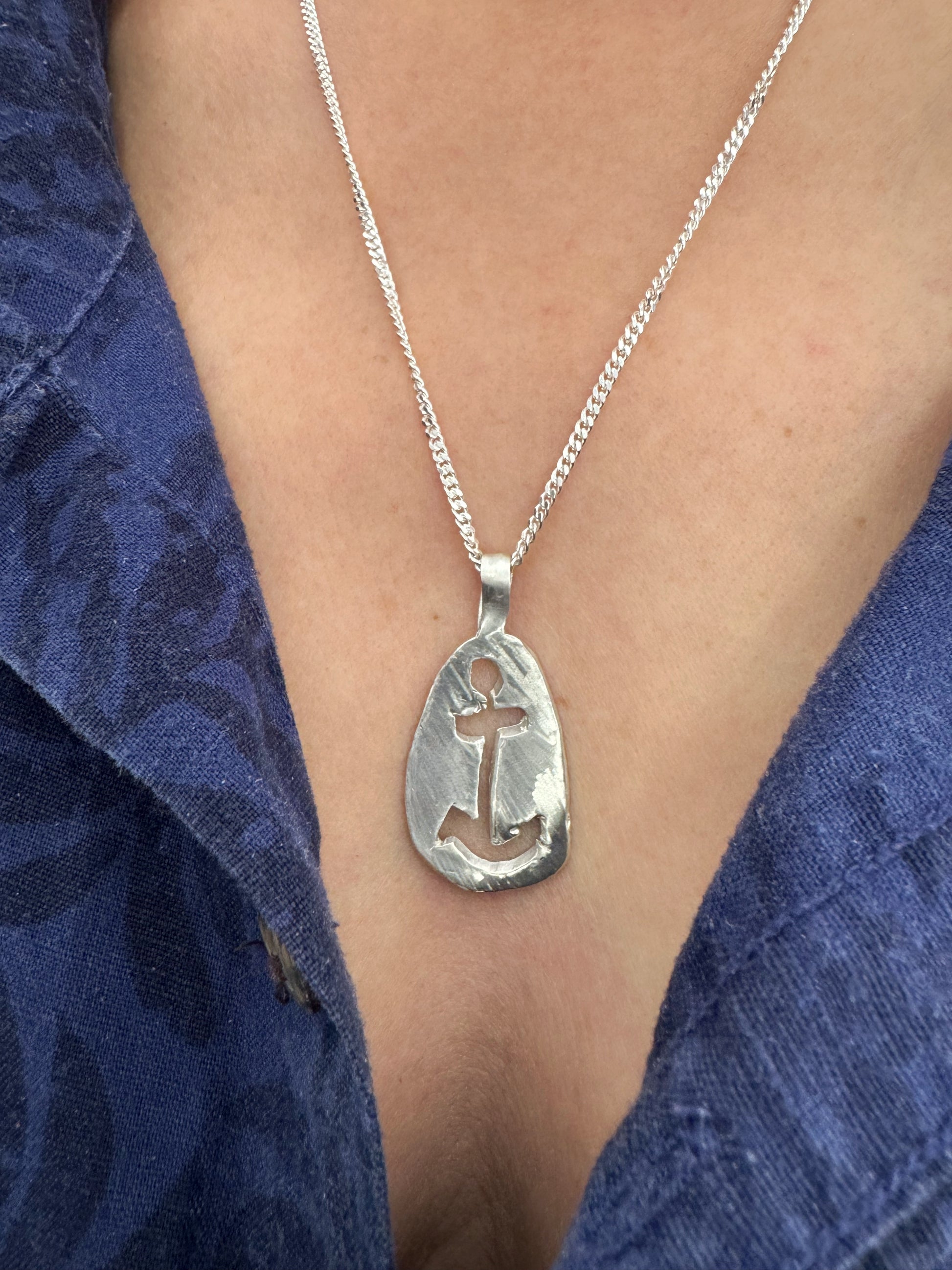 A person wearing the Anchor Sketch Pendant Inspired by Bernard Moitessier’s Spirit on a chain necklace, against the background of a blue fabric.