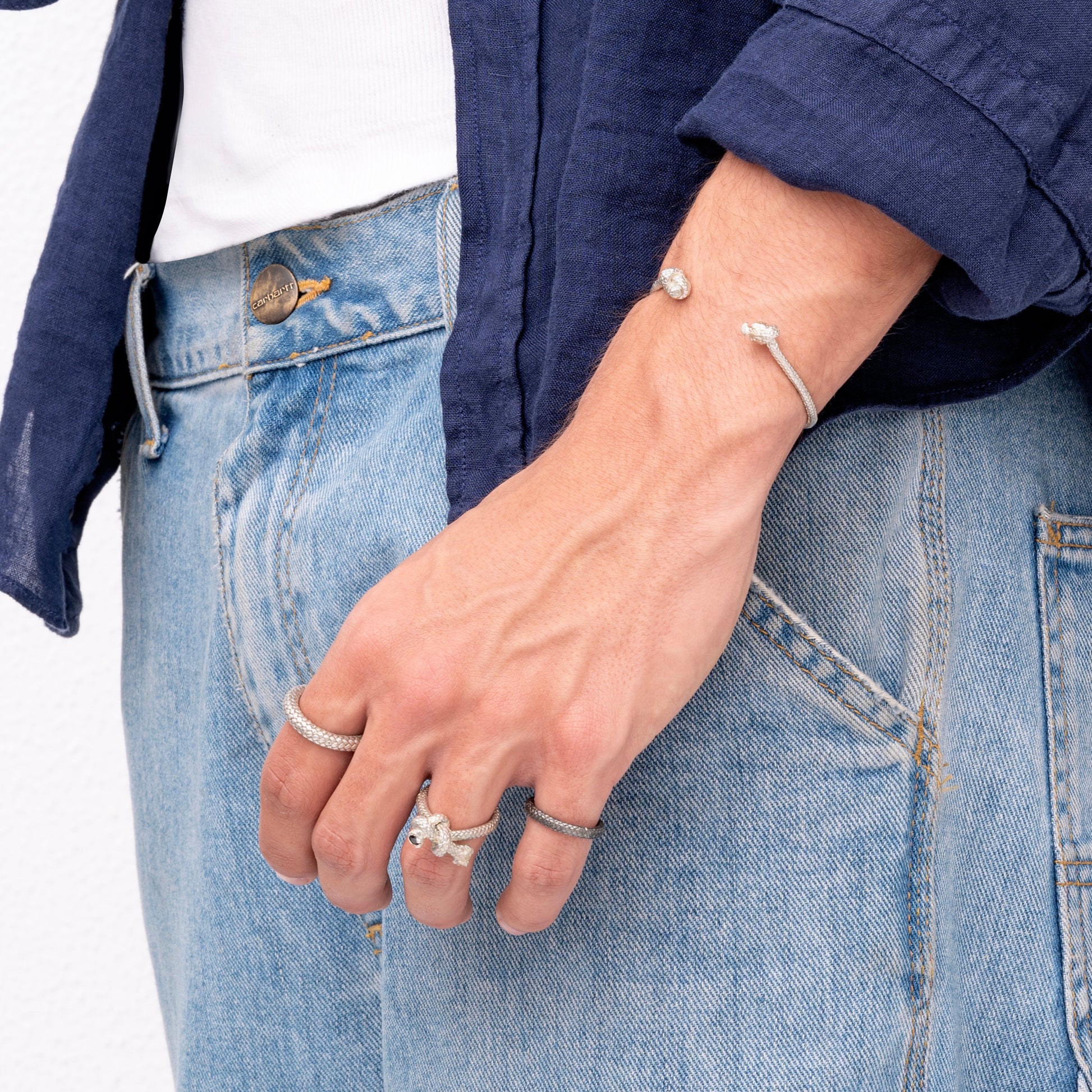 A person wearing a blue shirt and light blue jeans is displaying their hand adorned with multiple sterling silver rings and a Nautical Atlantic Bracelet Rope Cuff.