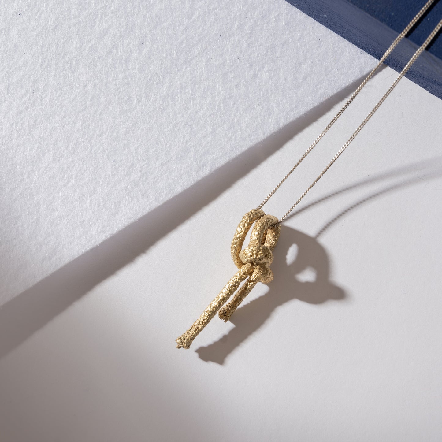 A Nautical golden knot pendant - Ballestrinque S hangs on a thin silver chain, casting a shadow on a white and blue surface.