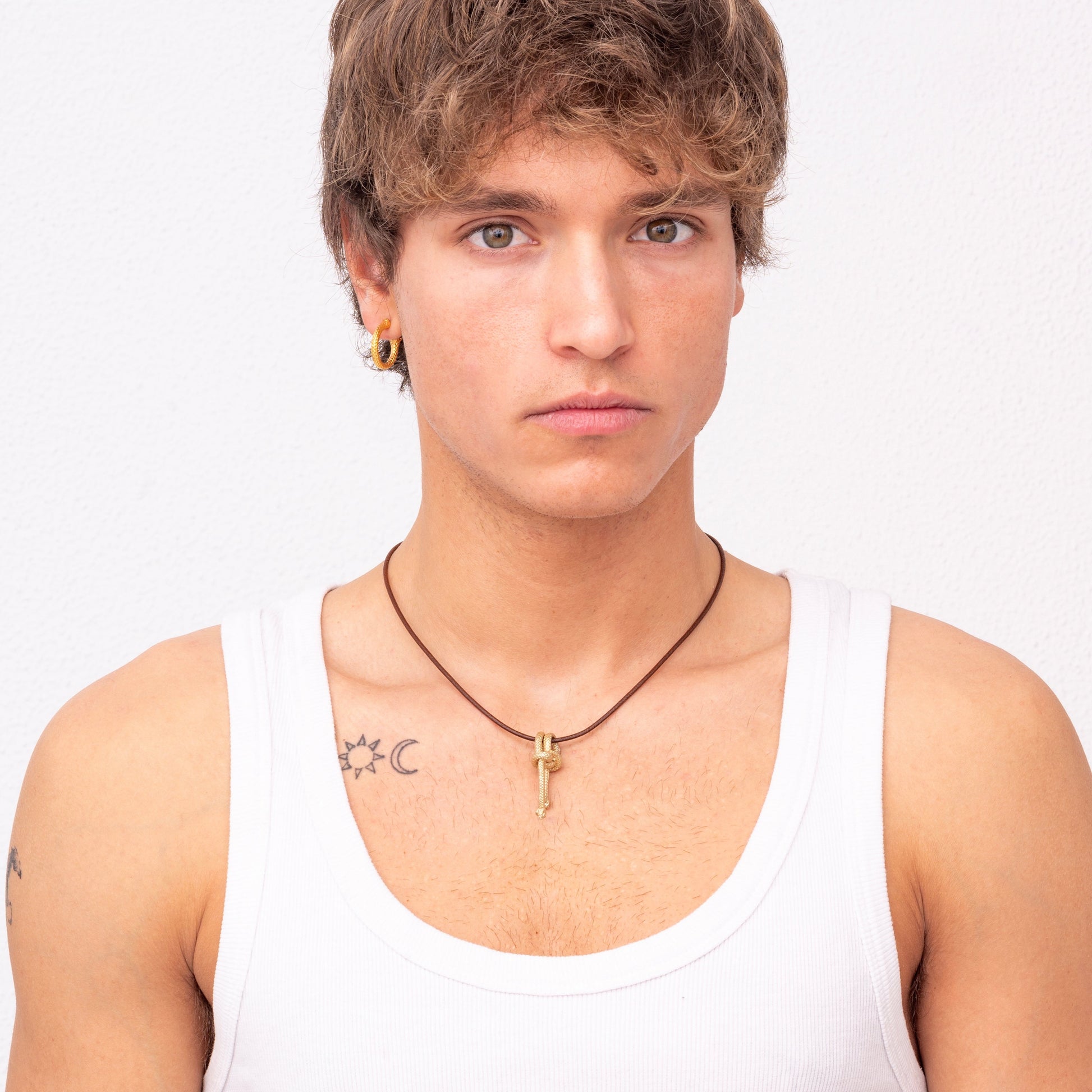 A person with short, light brown hair and blue eyes wears a white tank top, the Nautical Golden Knot Pendant - Ballestrinque S in green gold plated brass around their neck, and two earrings. Tattoos of a crescent moon and a sun are visible on their shoulders.