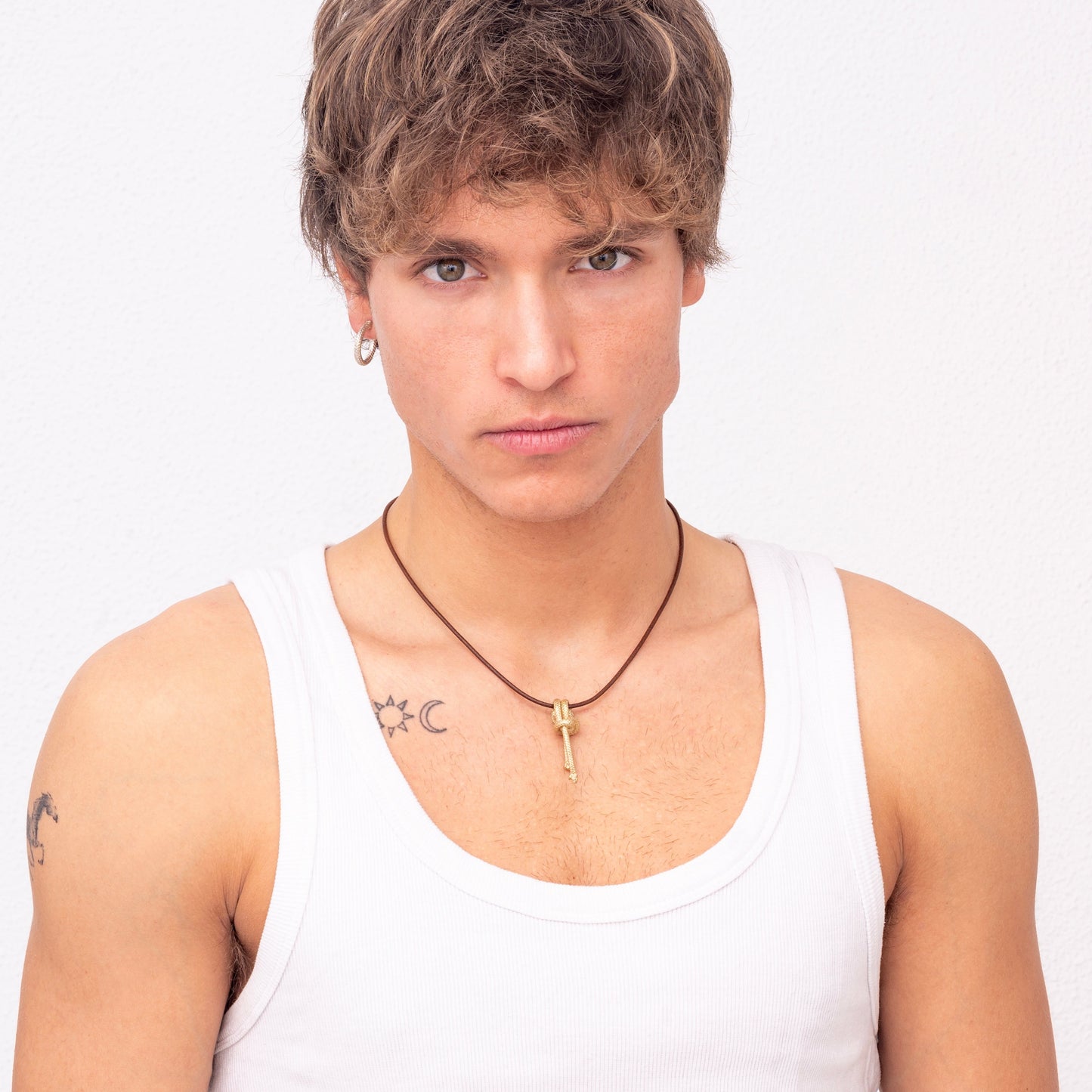 Young man with blonde hair wearing a white tank top, small hoop earrings, and a Nautical golden knot pendant - Ballestrinque S necklace, posing against a plain white backdrop. Has tattoos on both shoulders.