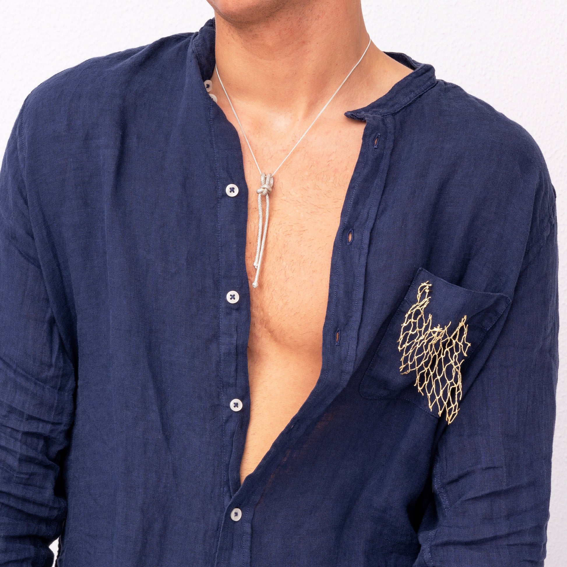 A person wearing an open dark blue shirt adorned with a gold chain detail on the pocket and a silver pendant necklace, exuding a sense of individuality reminiscent of the Handmade One of A Kind Ocean-Inspired Fishing Net Brooch.