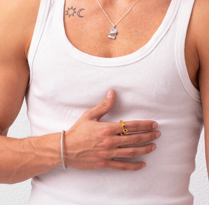 A person wearing a white tank top, a sterling silver necklace, a bracelet, and a Golden Ring Nautical Rope Double Sapphire touches their chest, revealing a sun and crescent moon tattoo on their upper chest.