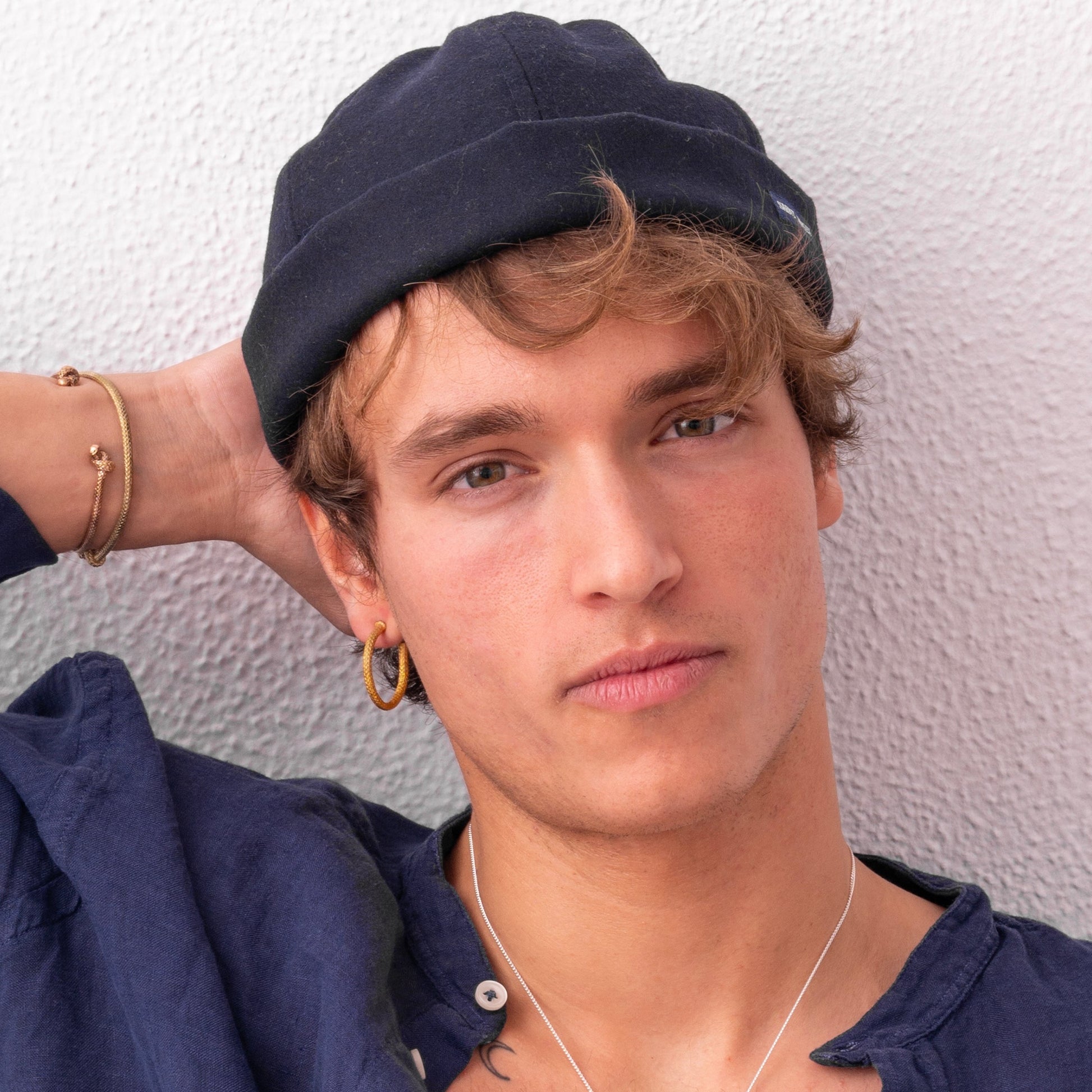 Person with wavy hair and a blue cap, wearing a blue shirt, and sporting the Nautical Hoop Corto Maltese Inspired: Creole Earring Rope Size M, looks at the camera with a neutral expression. One hand is resting behind their head.