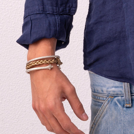 A person's wrist adorned with three bracelets: one braided, one beaded, and one Nautical Bracelet Torque Braid in Sterling Silver, wearing a blue long-sleeve shirt and light blue jeans.