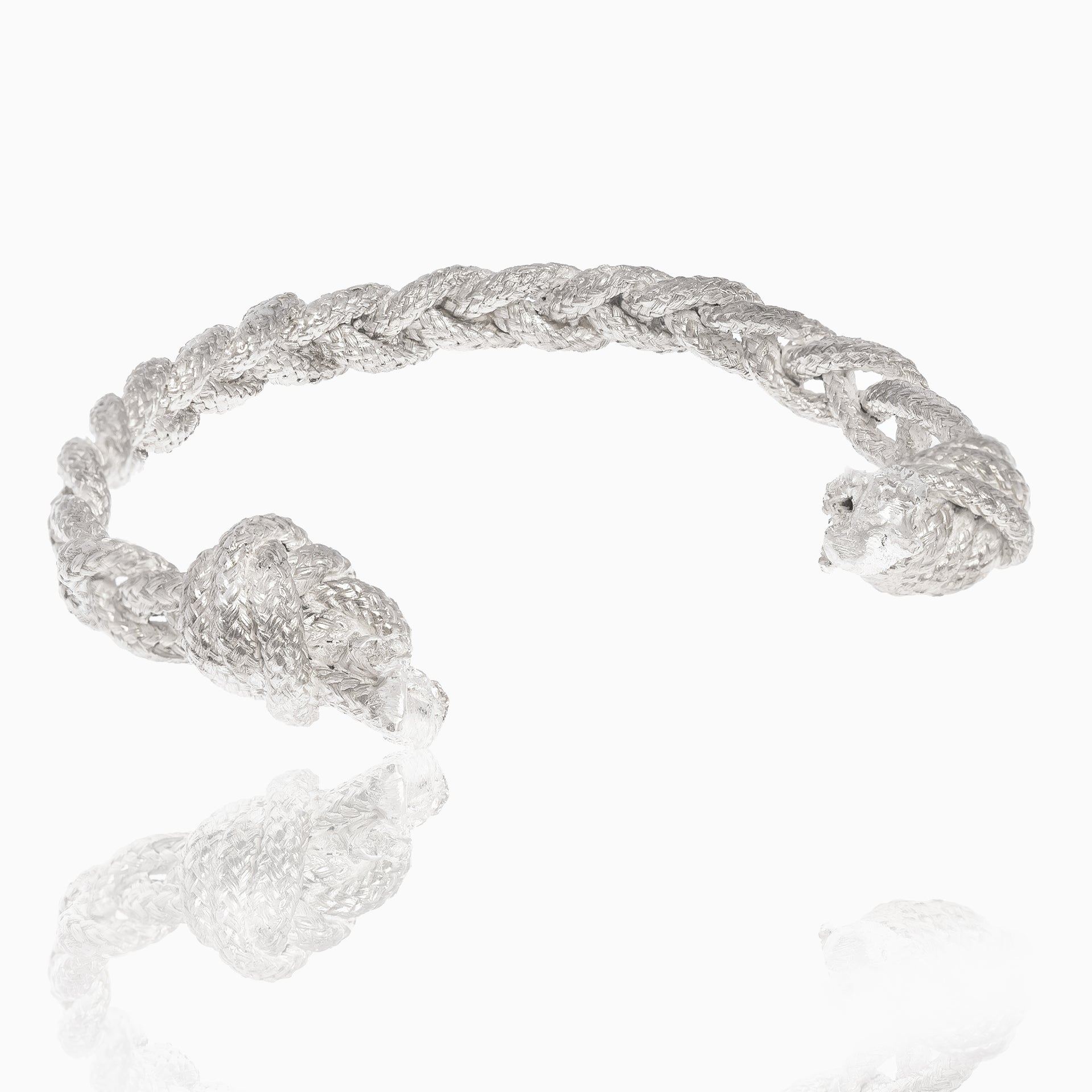 A handmade torque bracelet with intricate knot details on both ends, the Nautical Bracelet Torque Braid – available in sterling silver or brass – showcases exquisite craftsmanship, displayed on a reflective white surface.