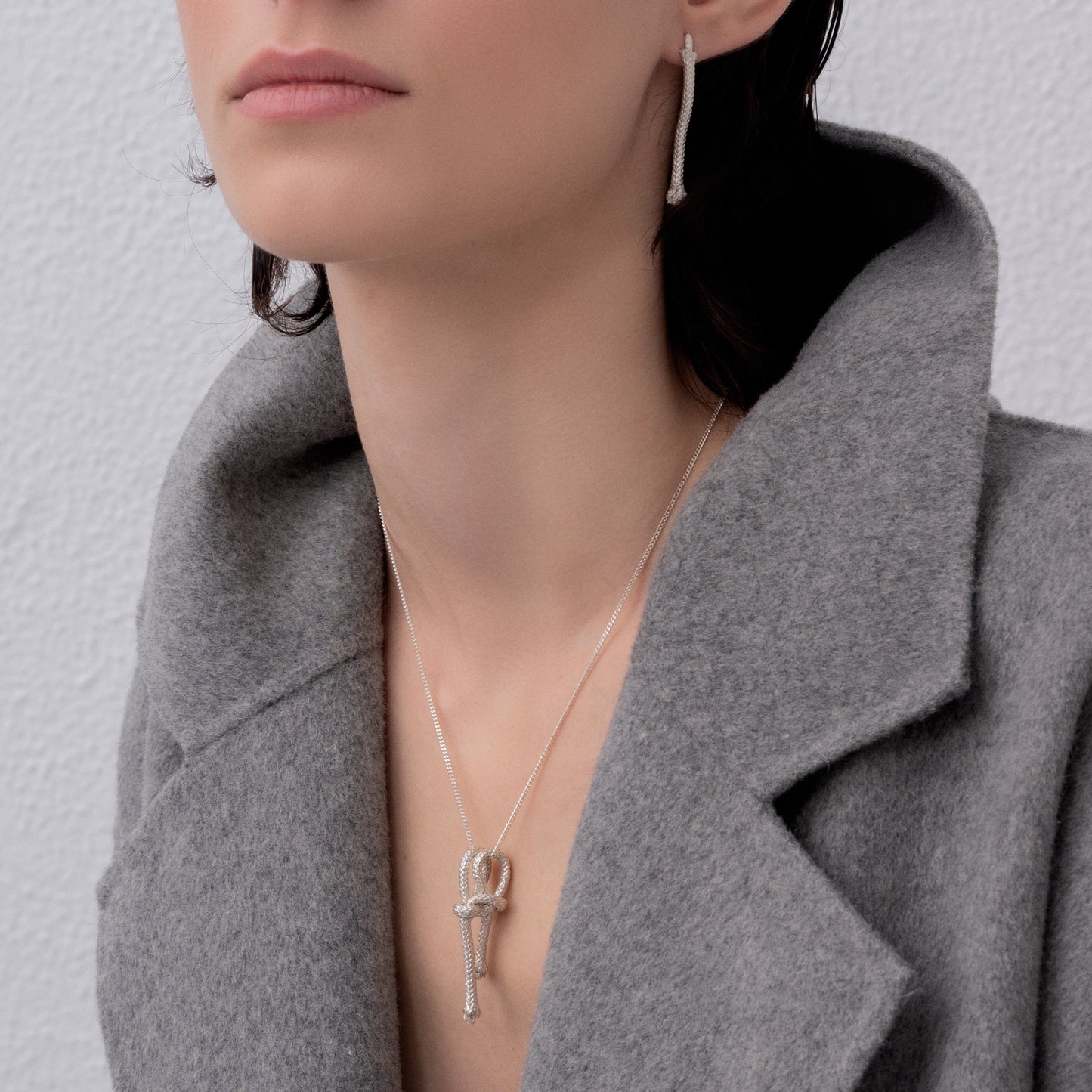 A person wearing silver jewelry, including the Ballestrinque Small Nautical Knot Pendant in Sterling Silver and a pair of rectangular drop earrings, paired with a light grey coat.