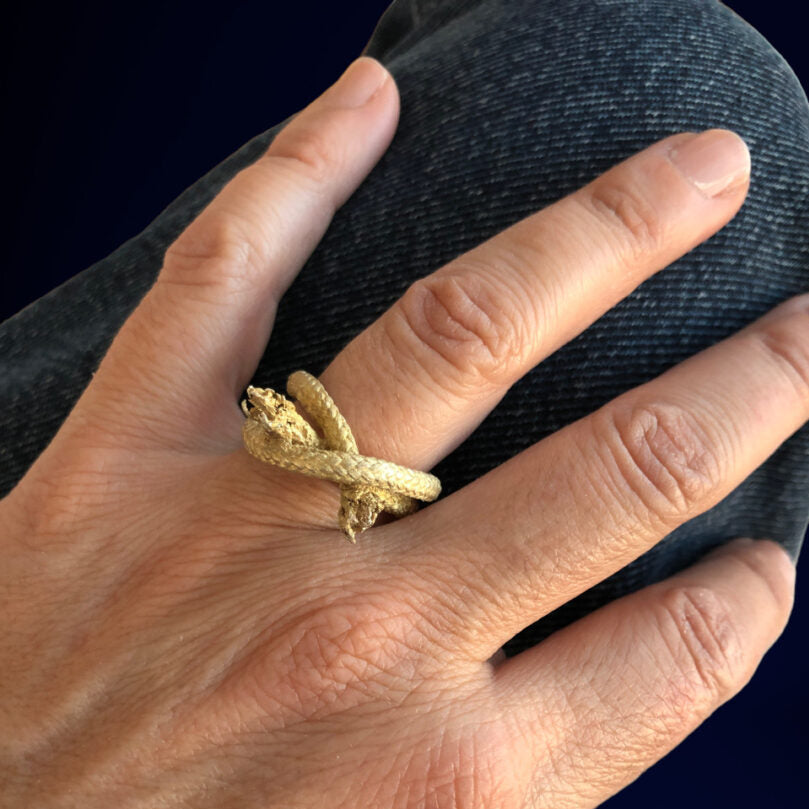 A hand is shown wearing the Ballestrinque Knot - Clove hitch - Artisanal Nautical Ring on the finger, with the name Angela Lago subtly etched inside. The hand rests on a denim-clad knee against a dark background.