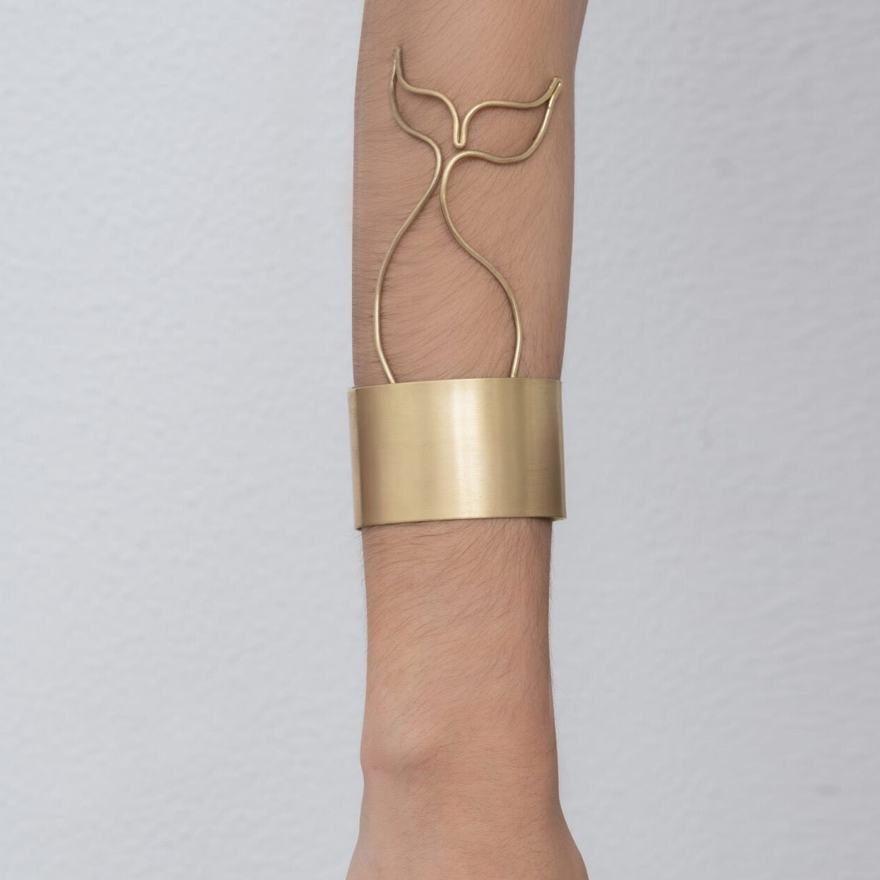 A person wearing a distinctive Whale Tail Cuff Bracelet Sailor's Tattoo Inspired on their forearm, featuring an abstract whale tail design and handmade by Angela Lago from 24k gold plated brass.