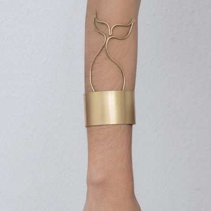 A person wearing a distinctive Whale Tail Cuff Bracelet Sailor's Tattoo Inspired on their forearm, featuring an abstract whale tail design and handmade by Angela Lago from 24k gold plated brass.