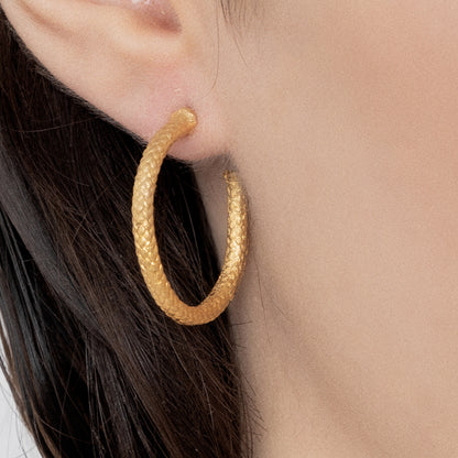 Close-up of an ear adorned with a textured, handcrafted Nautical Hoop Corto Maltese Inspired: Creole Earring Rope Size M. The person's light skin and dark hair are visible next to the ear, accentuating the exquisite detail of the gold hoop earring.