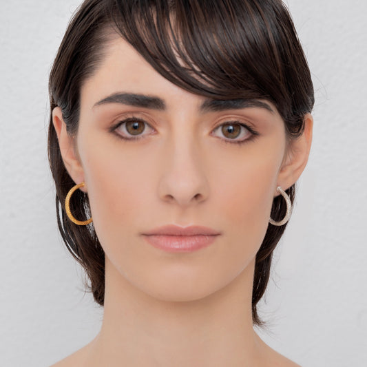 Close-up of a person's face with short dark hair, wearing Nautical Hoop Corto Maltese Inspired: Creole Earring Rope Size M sterling silver hoop earrings. The background is light grey.