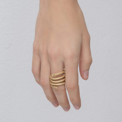 A hand adorned with the Curly Handcrafted Rope Ring III, a gold-plated brass (5 microns gold) masterpiece featuring an intricate, multi-band design that elegantly wraps around the finger. The plain, light-colored background accentuates the exquisite craftsmanship of Angela Lago's creation.