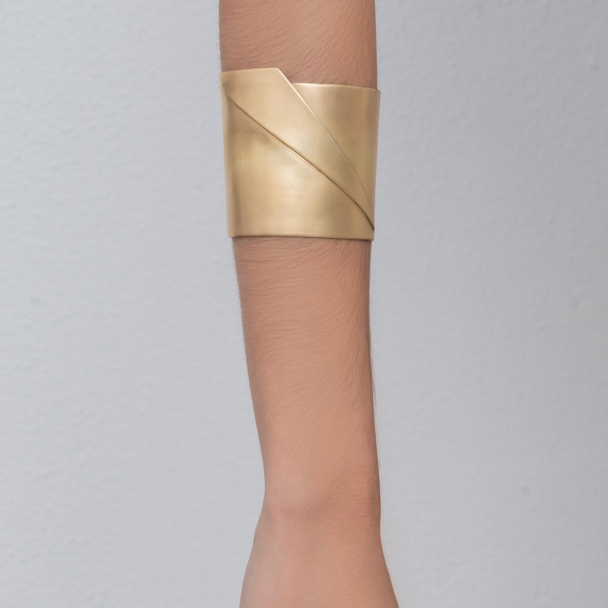 Close-up of a person wearing the Sea Wake bracelet, a wide, gold metallic cuff hand-made by Angela Lago, on their wrist against a plain, light gray background.