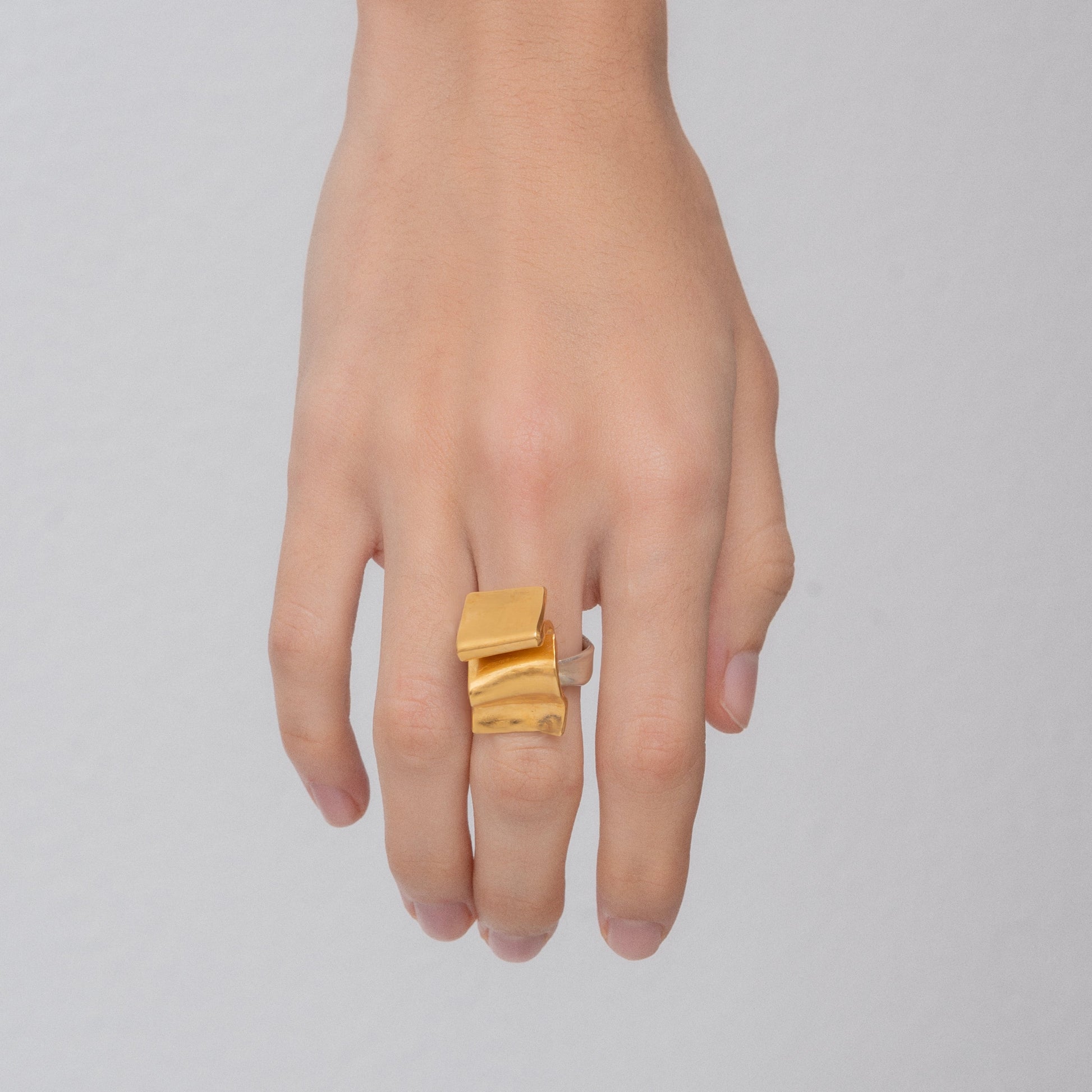 A hand wearing a handcrafted ring with a wave design, 24k gold-plated brass, on the middle finger against a plain background.