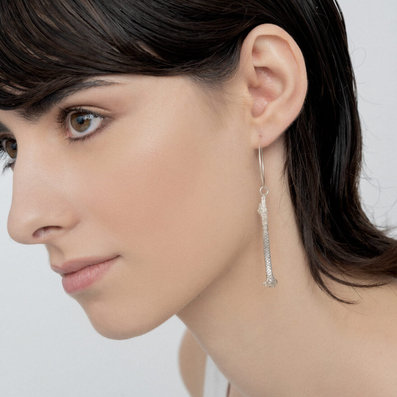 A person with short dark hair is wearing a long, artisan-made Thin Hoop Nautical Earring with Sterling Silver Rope and looking towards the left. The background is plain and light-colored.