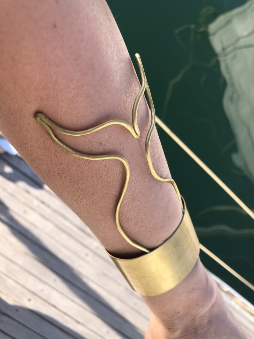 A handmade brass "Whale Tail Cuff Bracelet, Sailor's Tattoo Inspired" by Angela Lago is elegantly worn on a person's forearm, set against a backdrop of a wooden surface and water.