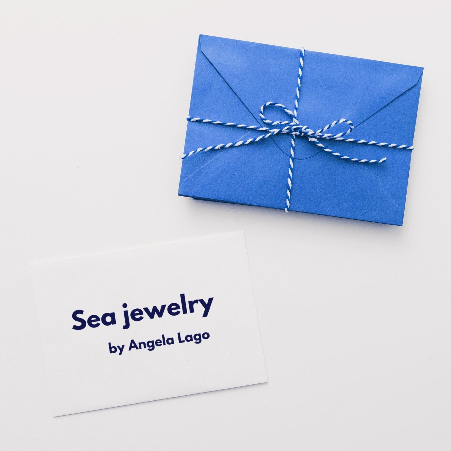 A white card that reads, "Angela Lago Sea Jewelry: Ocean-Inspired Treasures" sits next to a blue envelope tied with white and blue twine.