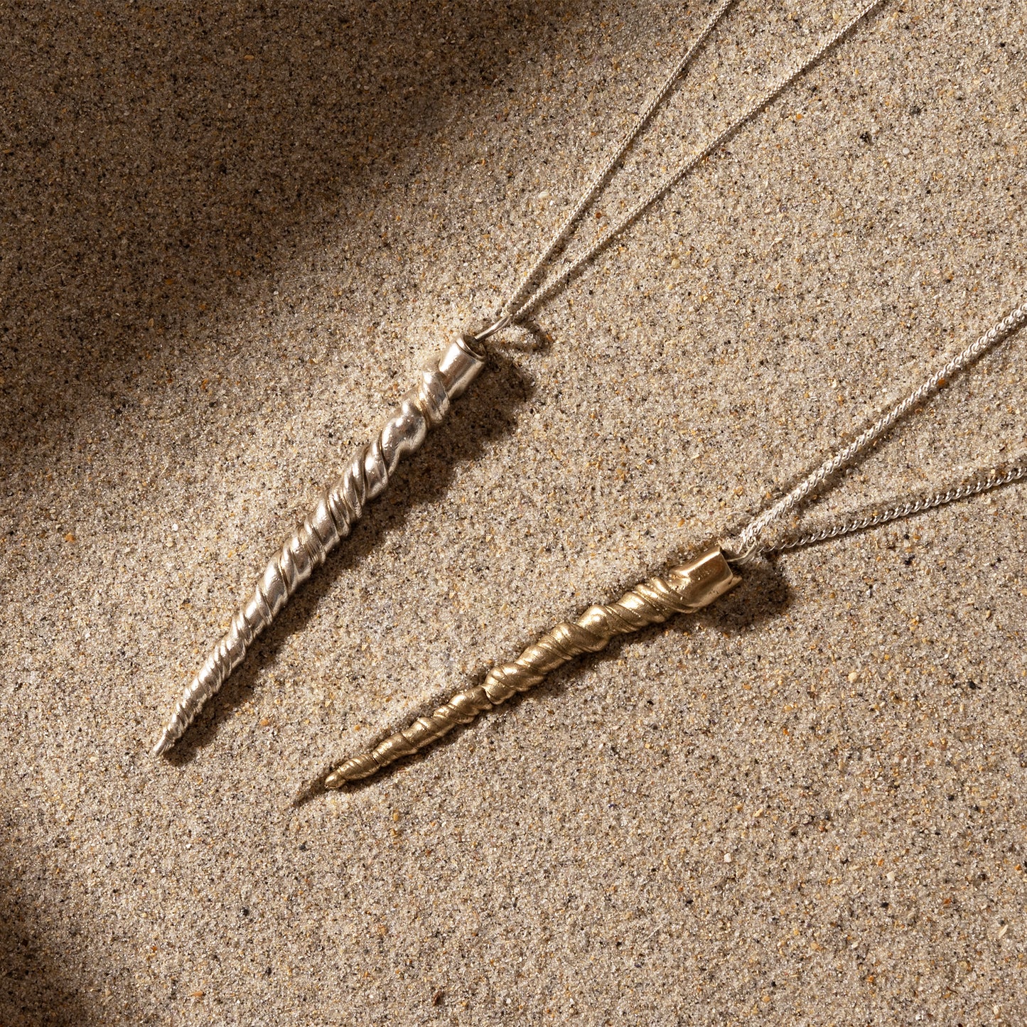 Two twisted metal pendant necklaces, one in sterling silver and the other in gold, lying on a sandy surface. These unique pieces resemble Narwhal Tusk Handmade Tooth Pendants and are crafted by Angela Lago.