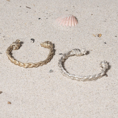 Two braided bracelets, one gold and one silver, rest on a sandy surface near a small seashell.