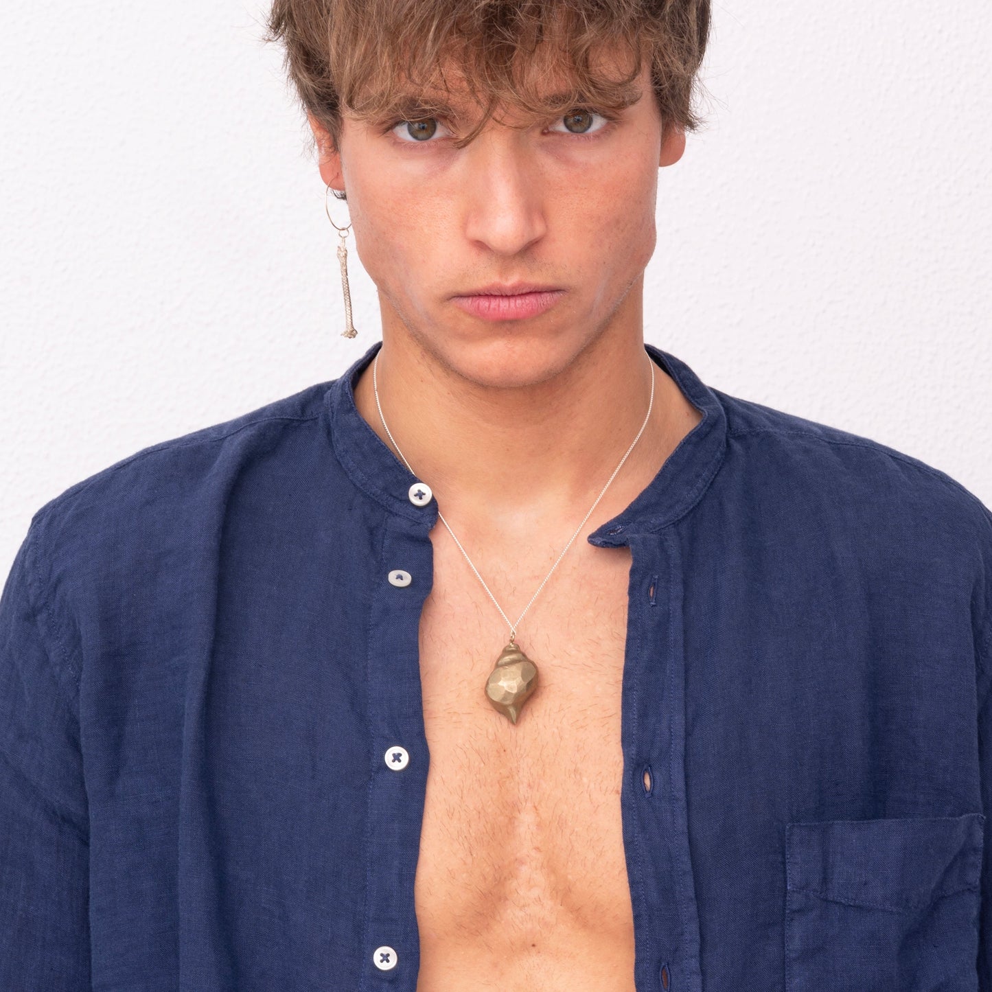 Young man with light brown hair wearing a navy blue shirt, a single earring, and the Nautical Handcrafted Seashell One of A Kind Pendant Nº 1 by Angela Lago poses against a plain white background.