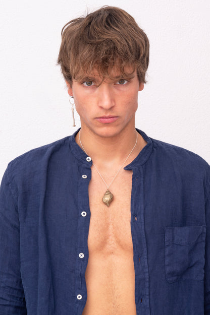 A person with short hair and a serious expression wears a blue unbuttoned shirt, an artisan-made necklace with a pendant, and a Thin Hoop Nautical Earring with Sterling Silver Rope, standing against a plain white background.