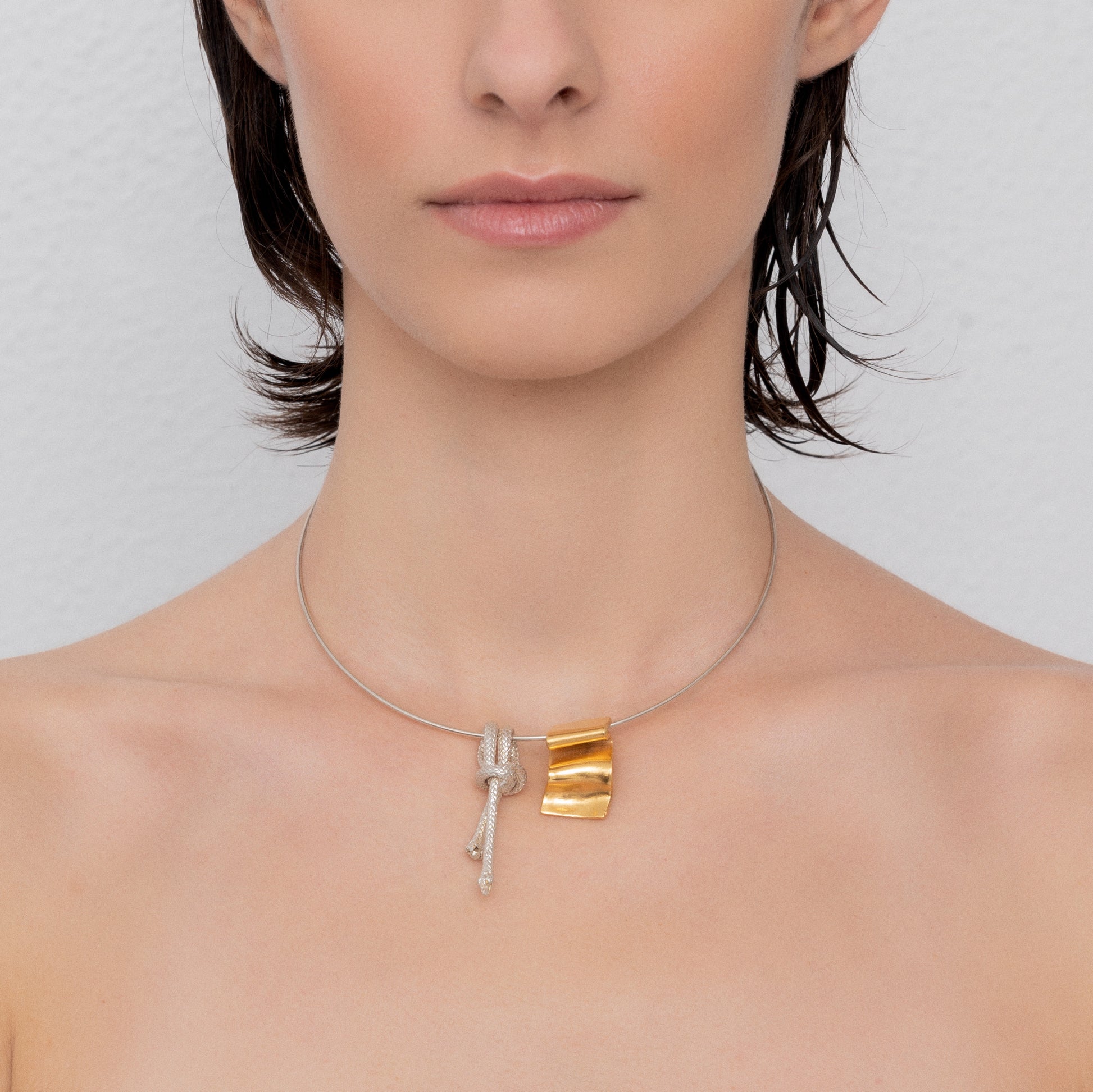 A close-up of a person wearing the Ballestrinque Small Nautical Knot Pendant in sterling silver, featuring a minimalist Clove hitch knot and a rectangular piece in metallic tones, hanging from a sterling silver chain. The background is plain.