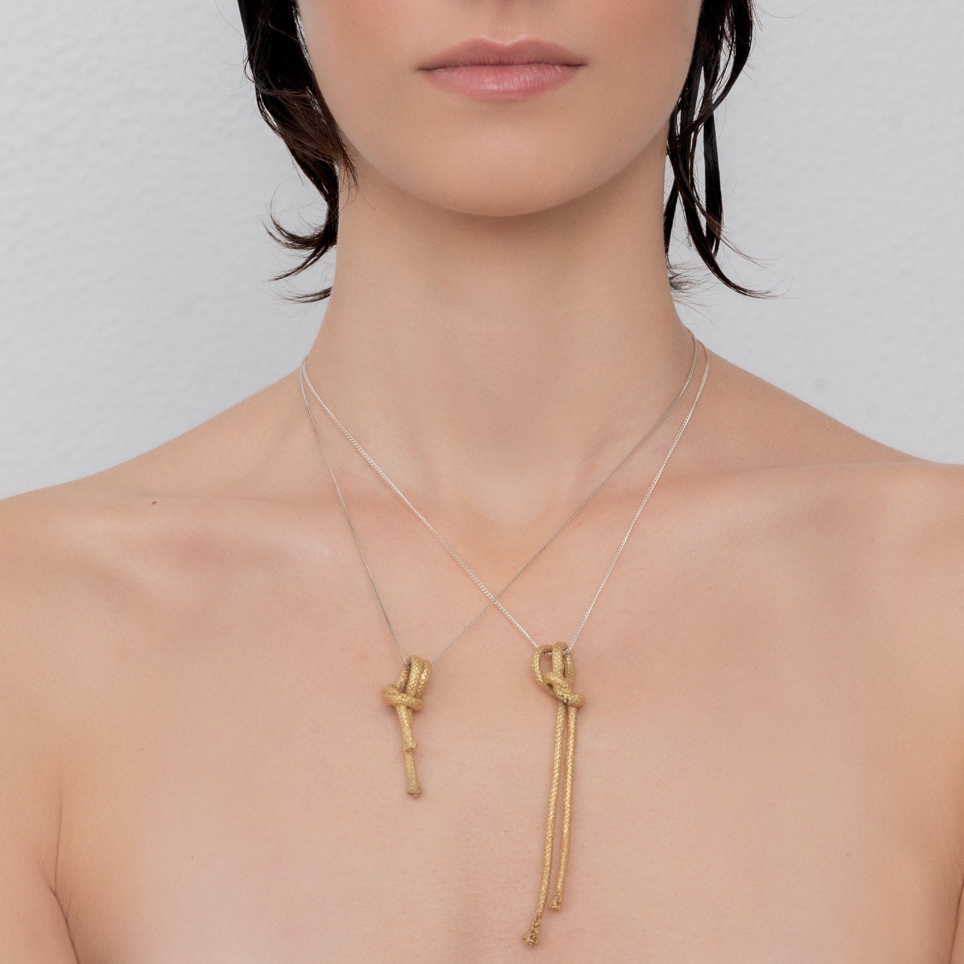 Close-up of a person wearing two green gold plated brass 'Nautical golden knot pendant - Ballestrinque S' necklaces, one shorter and one longer, against a plain background.