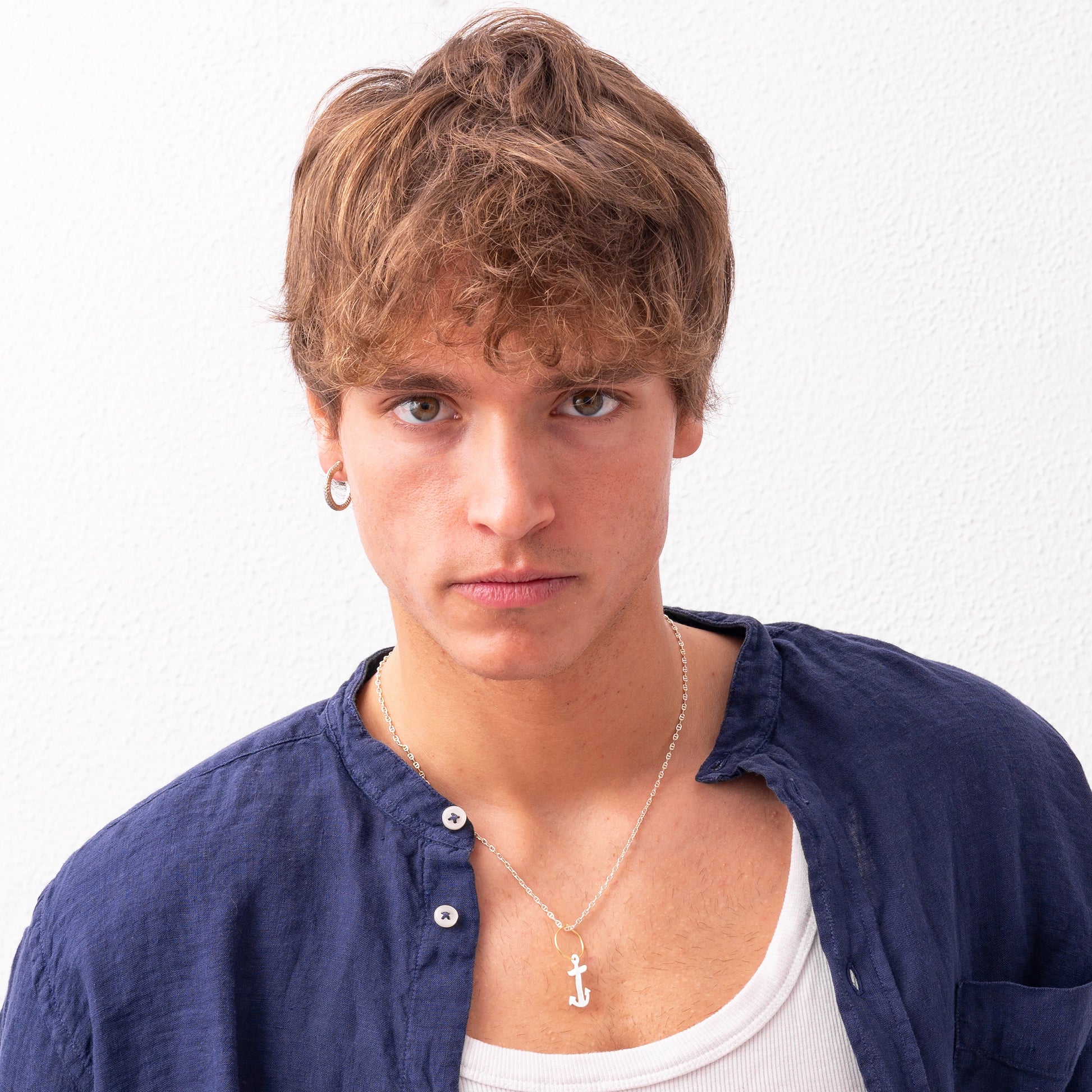 A young person with curly hair wears a blue shirt over a white tank top and a necklace with an anchor pendant, sporting the Nautical hoop Corto Maltese inspired: Creole Rope Earring size S, handmade by Angela Lago, while posing against a white background.