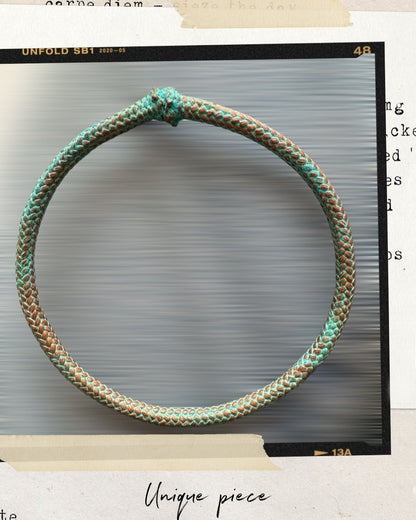 A circular, intricately woven bangle with green and brown threads is centered against a blurred background. The border appears like a vintage photograph with "Unique piece" at the bottom, showcasing the artisan-made elegance of the Artisanal Bronze Rope Bangle Inspired by Natural Nautical Oxidation.