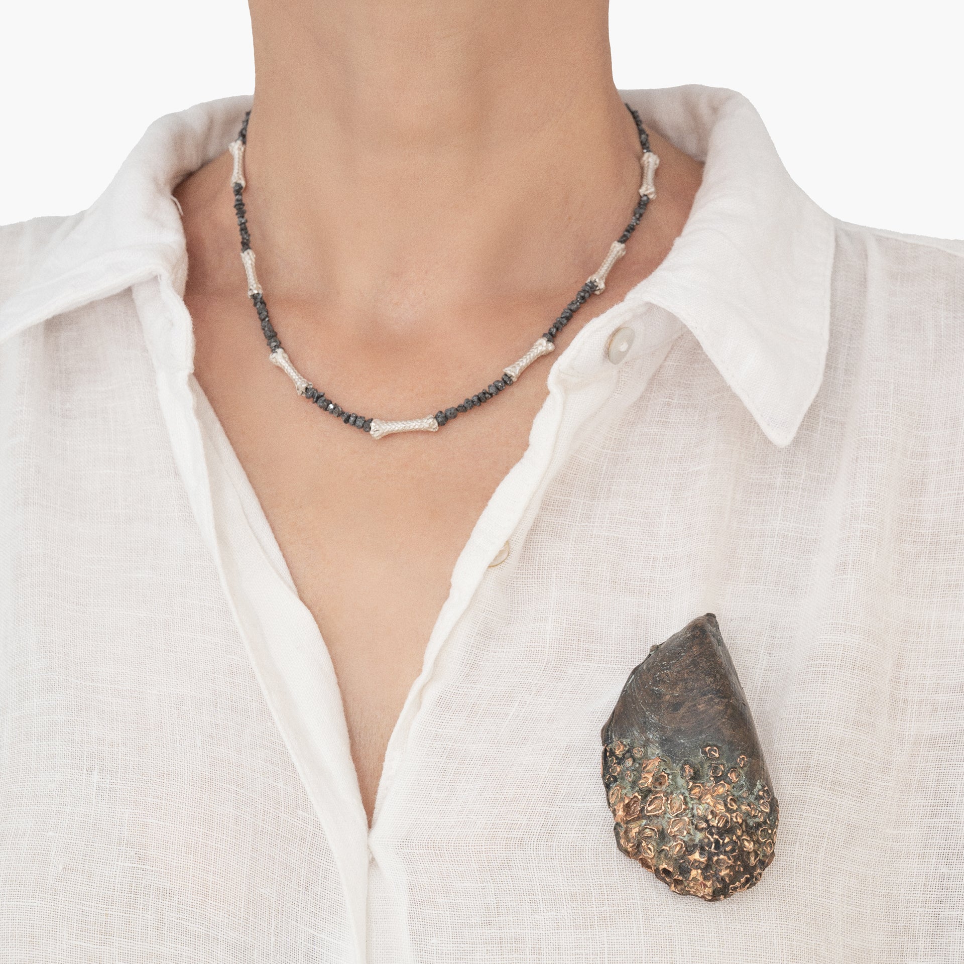 A person wearing a white shirt features a necklace made of beads and knots, along with a Bronze Mussel Brooch with Patina – Unique Piece, which resembles a textured stone with metallic elements.