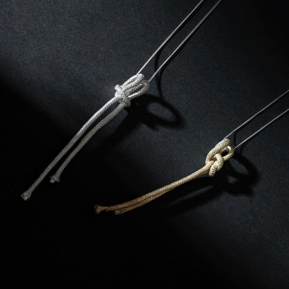 Against a dark background, two ropes are displayed – one in gray and the other in tan – each intricately tied in different types of knots. The Artisanal Made Ballestrinque Nautical Knot Pendant adds an elegant touch to the scene, evoking a nautical theme that's perfect for a sterling silver pendant collection.