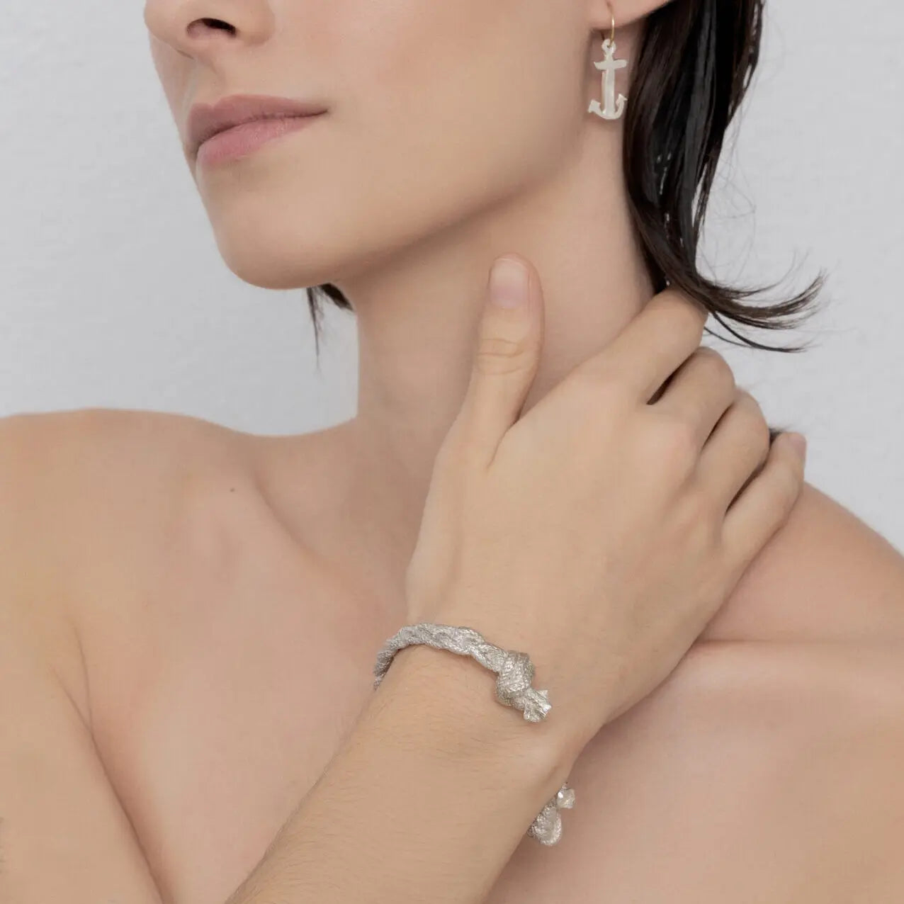 A person is shown wearing the Nautical Bracelet Torque Braid with braided details, paired with sterling silver anchor-shaped earrings. Their hand is gently touching their neck.