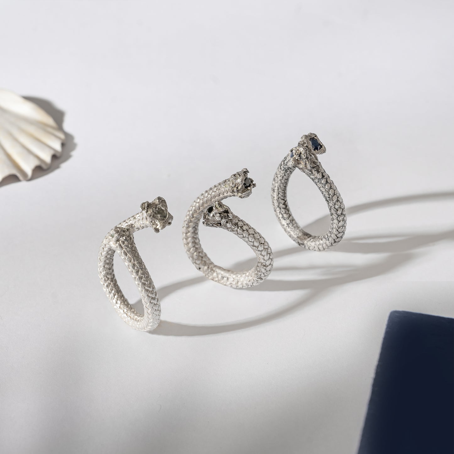 Three Aquamarine Artisanal Rope Rings in Sterling Silver are elegantly displayed on a white surface. A seashell and part of a navy blue object are partially visible on the sides, adding to the serene composition.