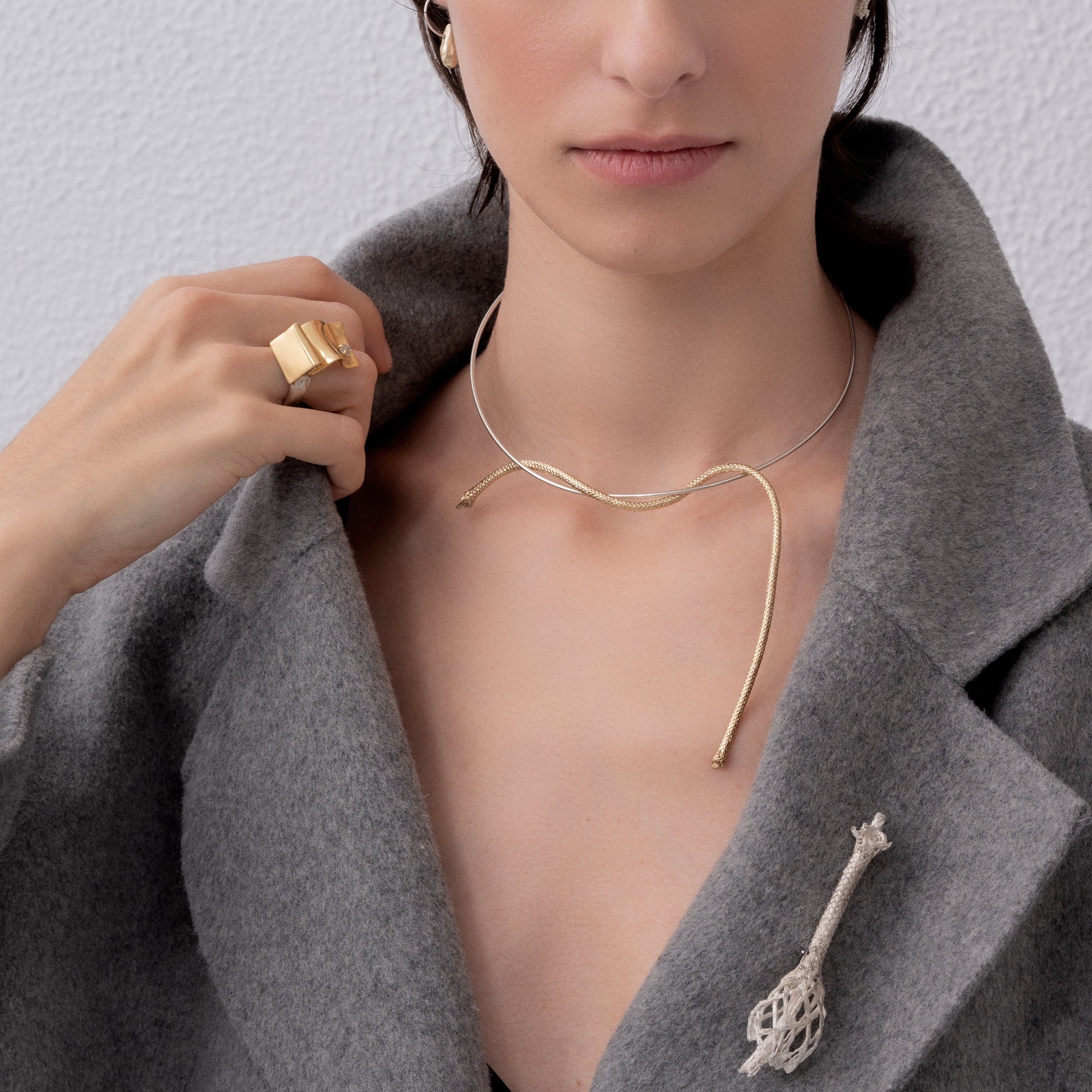 A person in a grey coat showcases a gold ring, an artisanal Choker Loose End Nautical Rope necklace, and a brooch shaped like a root or branch.