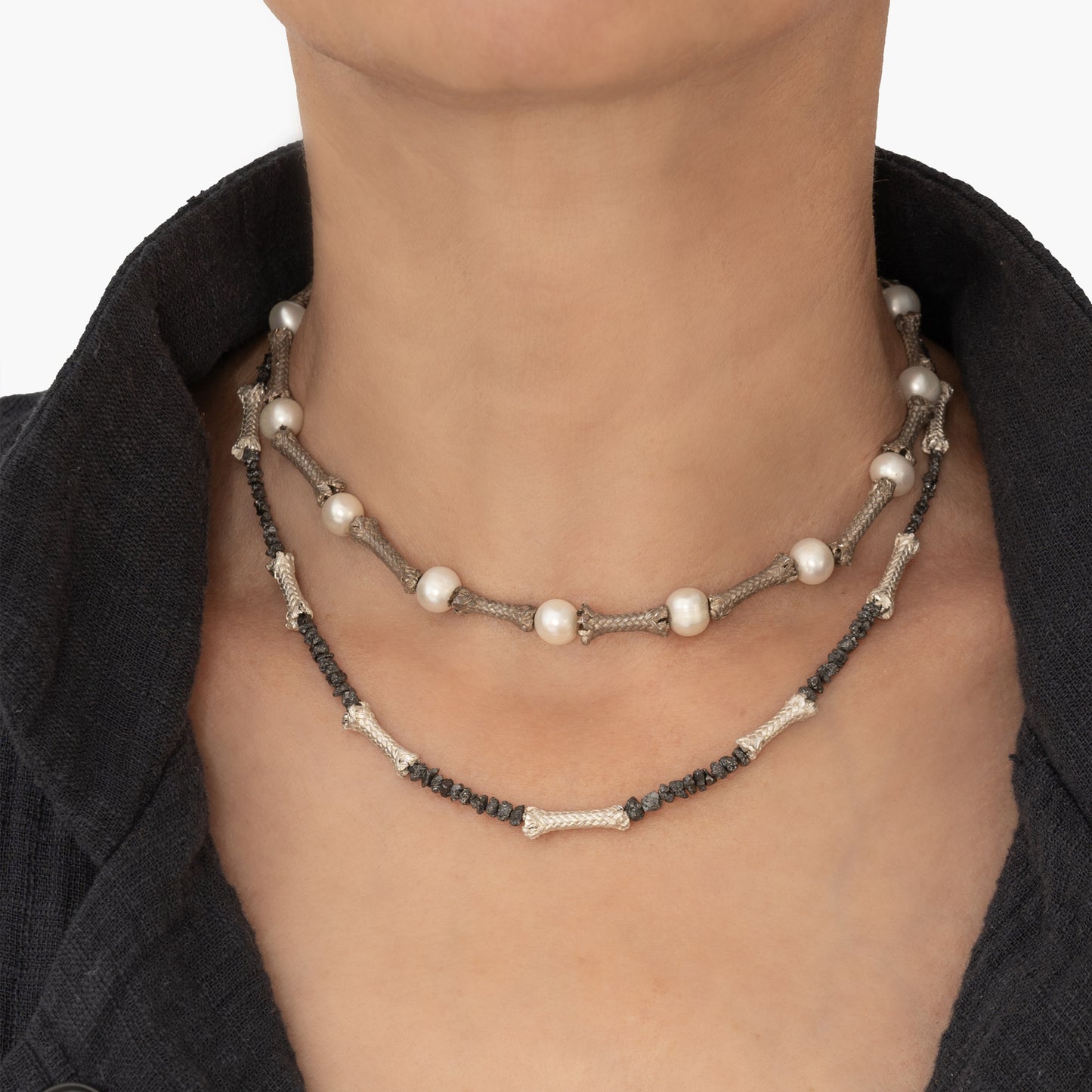 Close-up of a person's neck adorned with two stone bead necklaces: one featuring larger white beads and another showcasing smaller dark beads, both intricately designed with Raw black diamonds and Sterling silver ropes, complementing the dark jacket collar visible.