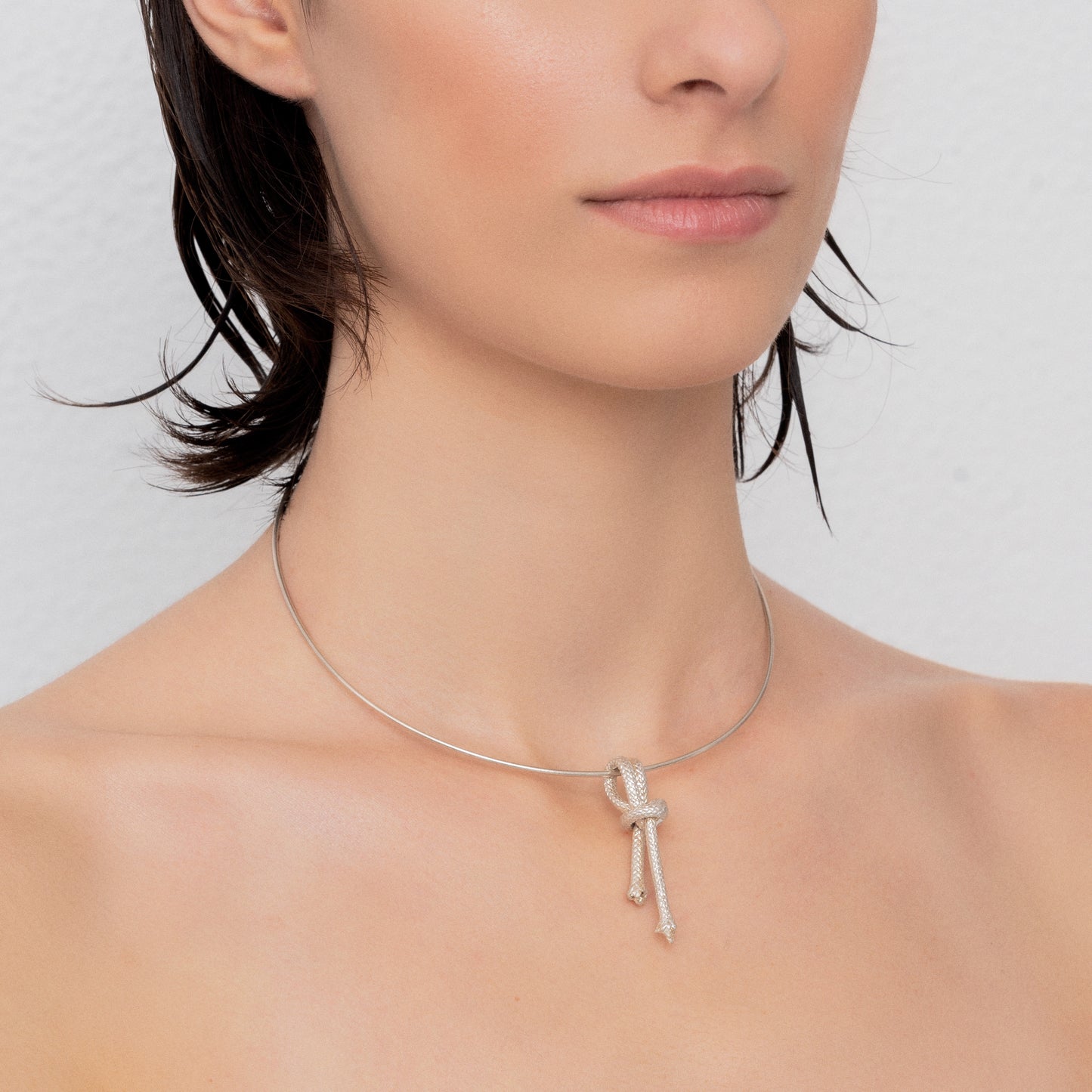 A close-up of a person wearing the Ballestrinque Small Nautical Knot Pendant in Sterling Silver. The pendant, featuring a delicate clove hitch knot, rests at the center of the sterling silver chain. The person's head is slightly turned to the side, and they have short, dark hair.